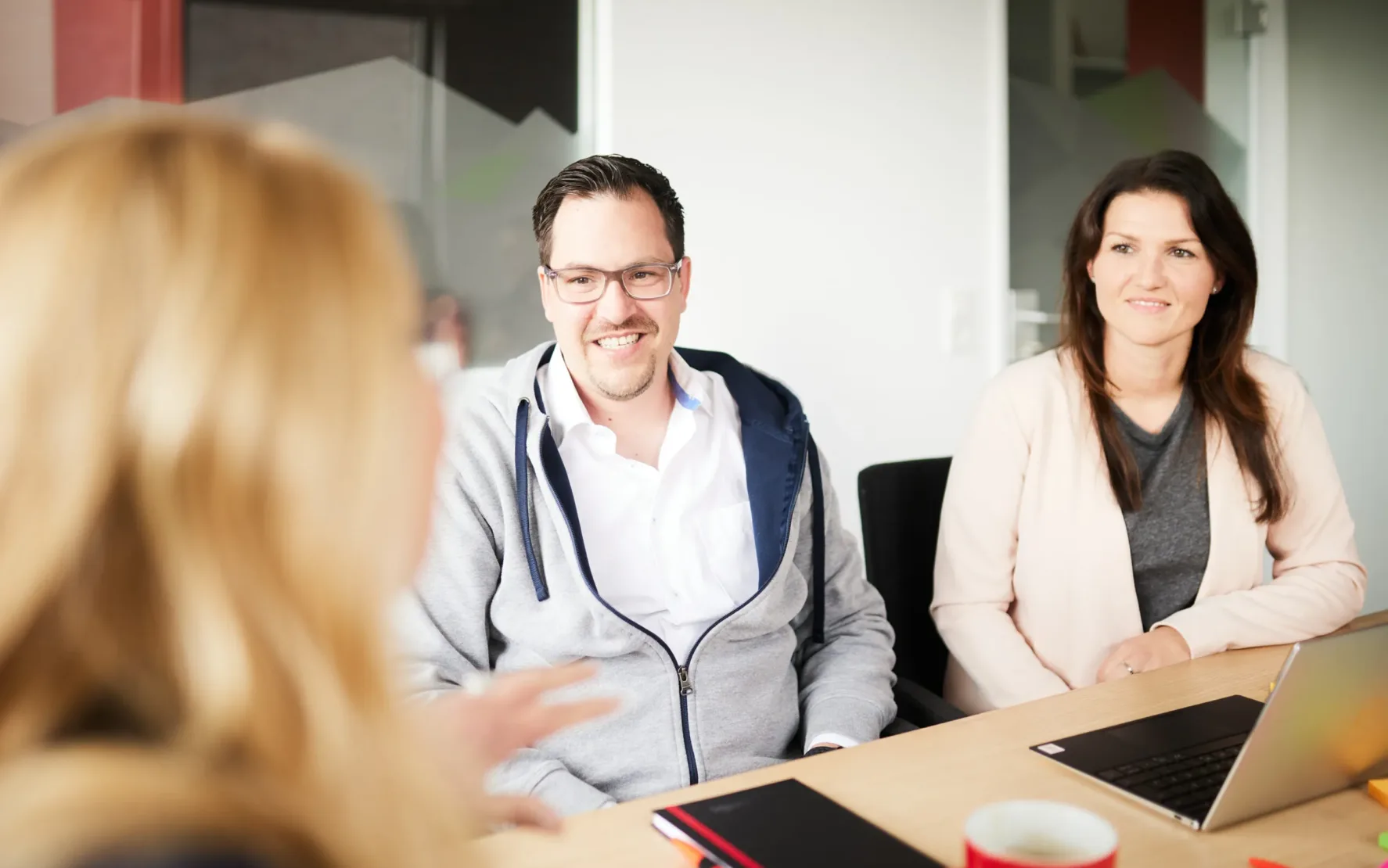 Foto von einem Mann und einer Frau, die an einem Konferenztisch sitzen und sich von einer Dame beraten lassen.