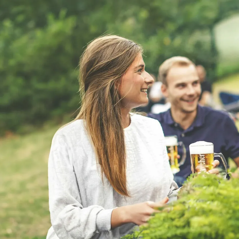 krombacher-freunde-seiten-stage-visual-freunde Eine Frau und ein Mann, beide mit Bierkrügen in der Hand, lächeln und unterhalten sich im Freien. Andere Leute im Hintergrund genießen ebenfalls ein Beisammensein auf einer Rasenfläche.